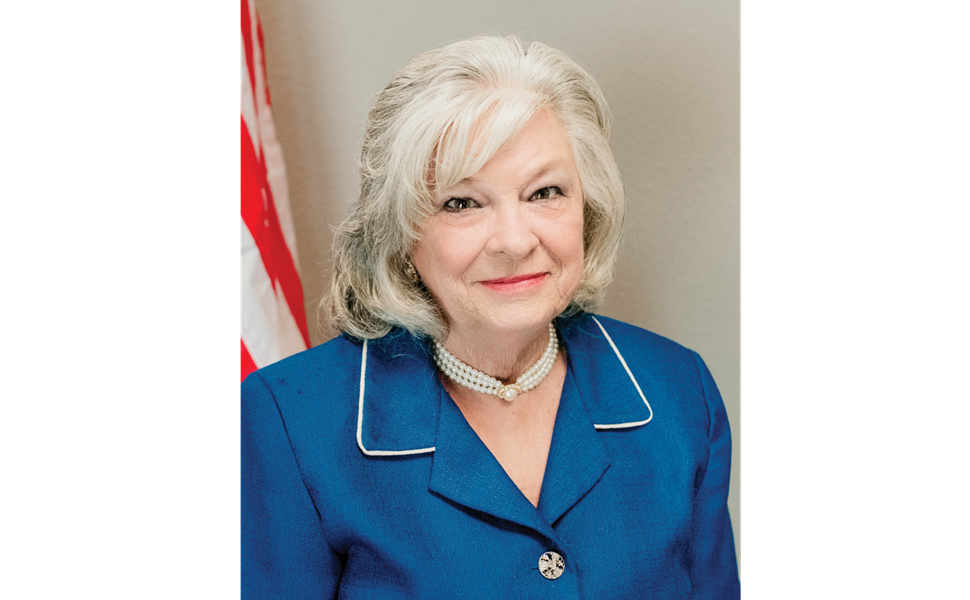 Official portrait of former Wilmer Mayor Shiela Petta in a blue blazer with a pearl necklace, standing before an American flag